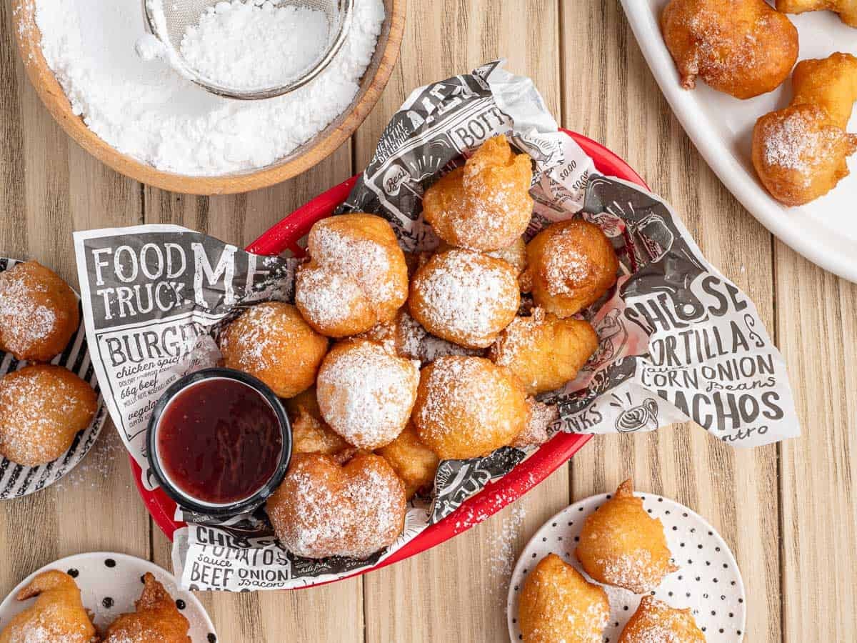 A basket of golden, powdered sugar-dusted beignets sits on a lined paper, reminiscent of Funnel Cake Bites. A small cup of red dipping sauce and plates of more beignets with a bowl of powdered sugar rest nearby on the wooden table.