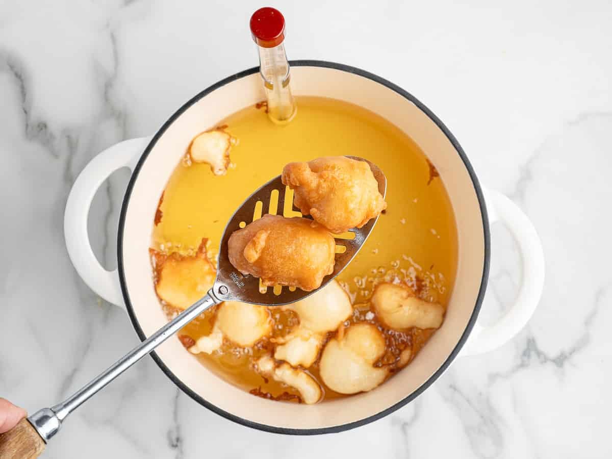 A hand holds a slotted spoon lifting two golden-brown Funnel Cake Bites from hot oil. Other bites are frying alongside, while a thermometer is clipped to the potโperfect for trying your favorite Funnel Cake Bites Recipe at home.