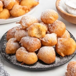 A plate of golden, round Funnel Cake Bites dusted with powdered sugar, with one bite broken open to show its fluffy inside. Bowls of chocolate sauce, jam, and powdered sugar are nearby—try this Funnel Cake Bites Recipe for a delightful treat.
