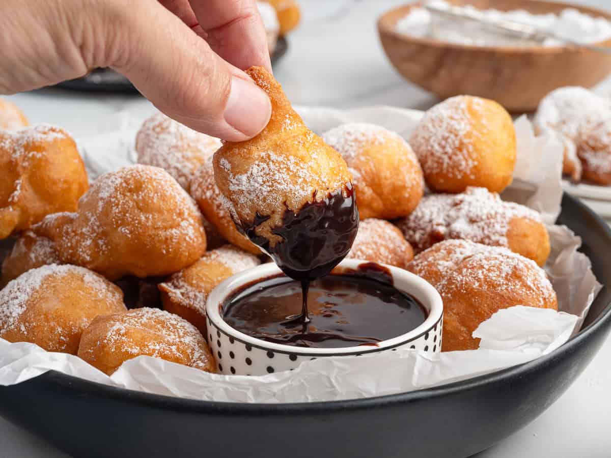 A hand dips a golden, powdered sugar-dusted Funnel Cake Bite into a bowl of chocolate sauce. These bite-sized treats are piled high on a plate lined with parchment paper, perfect for anyone craving this classic Funnel Cake Bites Recipe.