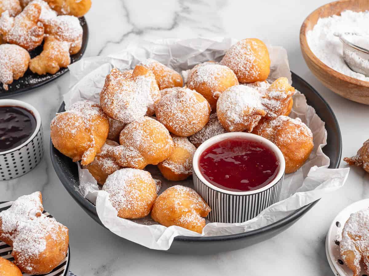 A plate of golden, powdered sugar-dusted beignets sits beside a bowl of red dipping sauce. Nearby, more treats and chocolate sauce temptโperfect inspiration for your next Funnel Cake Bites recipe.