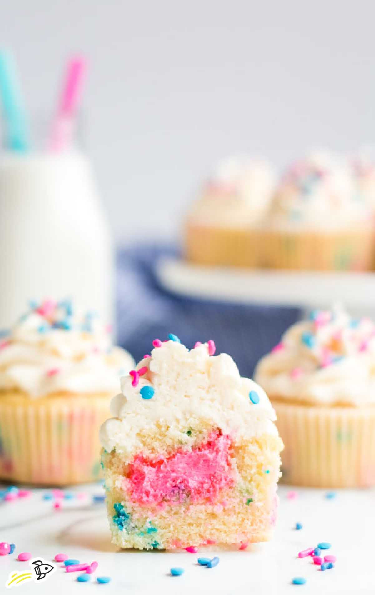 Funfetti-style gender reveal cupcakes with vanilla frosting and blue filling, surrounded by pink and blue sprinkles.