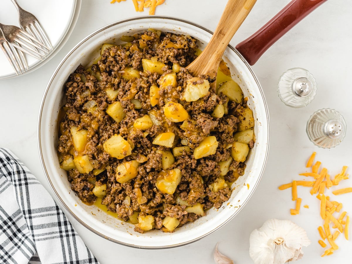 A skillet filled with cooked ground beef, diced potatoes, and melted cheese makes this Hamburger Hash Recipe irresistible. Shredded cheese, garlic, salt and pepper shakers, a checkered towel, and forks are nearby on a white surface.
