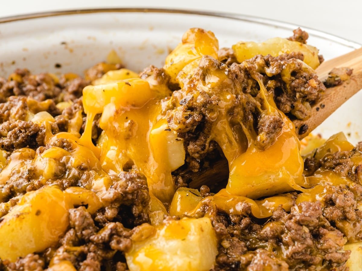 A close-up of a cheesy Hamburger Hash Recipe casserole, with melted cheddar cheese and ground beef mixed with diced potatoes, being scooped with a wooden spoon.