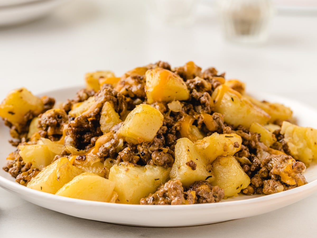 A white plate filled with a cooked mixture of ground beef and diced potatoes, seasoned and served on a light-colored surface. This hearty Hamburger Hash Recipe makes for a comforting and delicious meal.