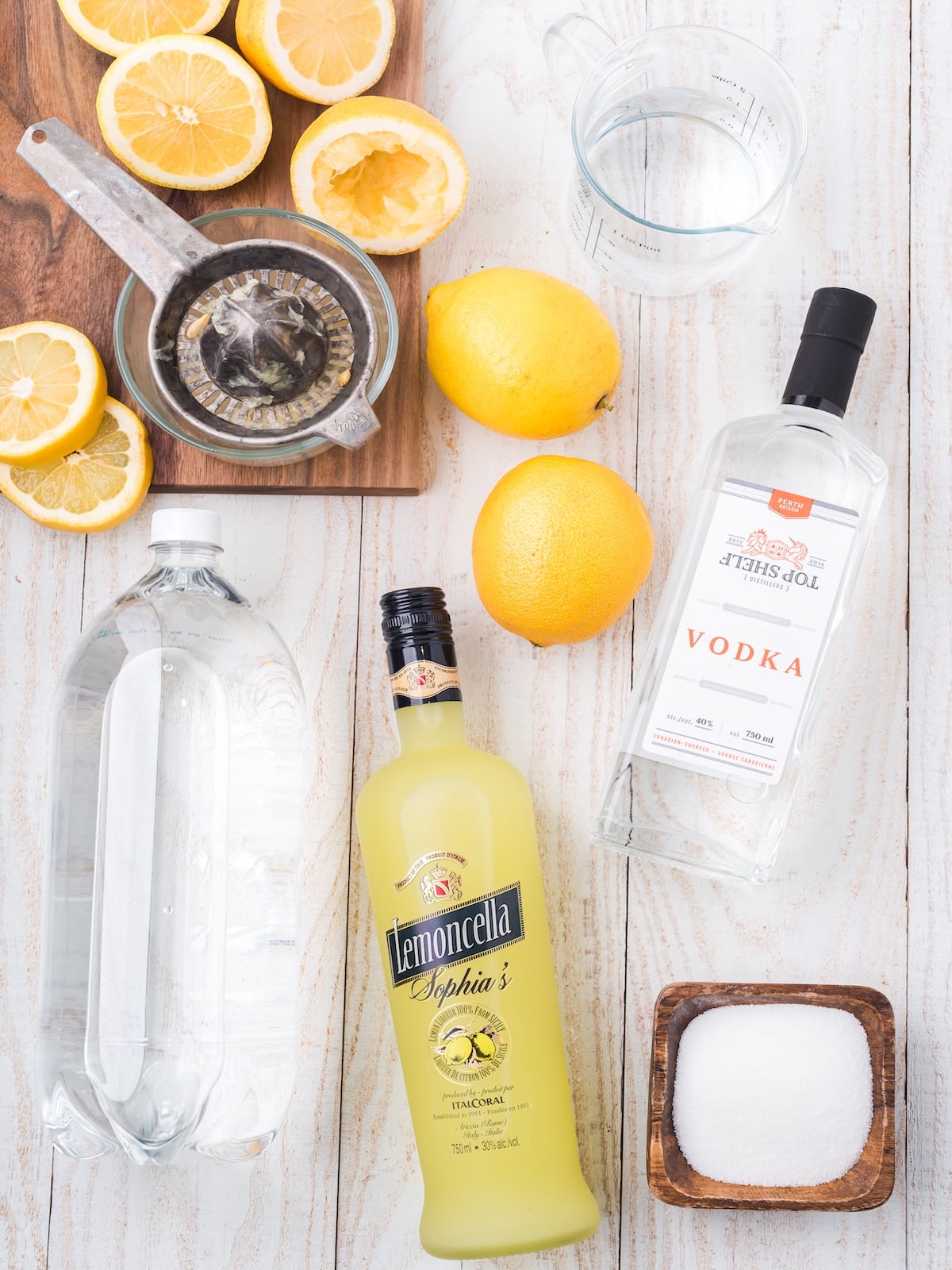 Overhead view of ingredients for a Hard Lemonade Recipe: halved lemons, lemon squeezer, two whole lemons, vodka, lemoncello, a pitcher and measuring cup of water, and a bowl of sugar on a white wooden surface.