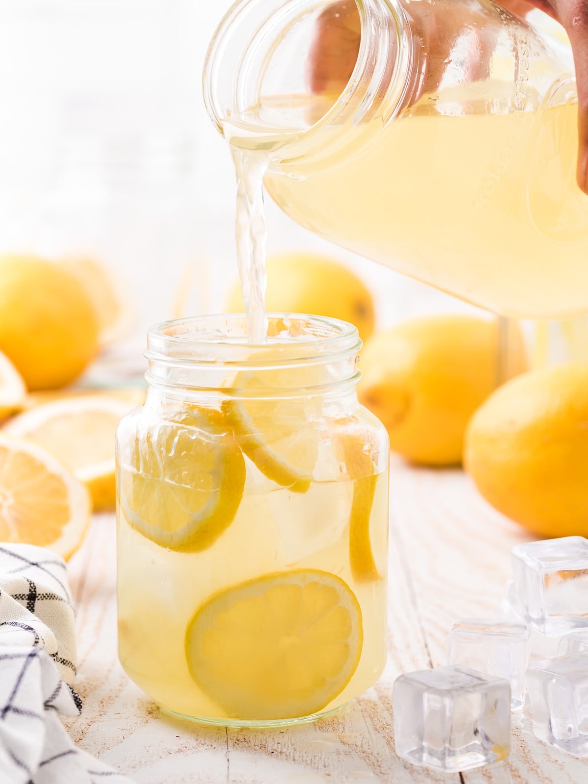 A hand pours hard lemonade from a glass jar into another jar filled with lemon slices. Fresh lemons and ice cubes are on a wooden table in the background.