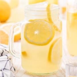 A mason jar filled with iced hard lemonade and lemon slices sits on a light surface beside a striped cloth, ice cubes, and whole lemons and lemon halves in the background&mdash;a perfect look for your next hard lemonade recipe.