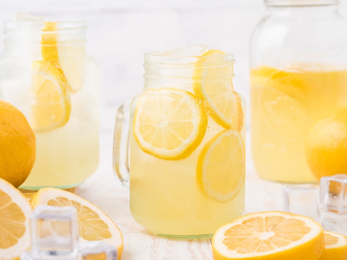 A mason jar filled with ice and Hard Lemonade, garnished with lemon slices, sits on a white surface. More Hard Lemonade in jars and pitchers, fresh lemons, and ice cubes surround the drink. The scene is bright and refreshing.