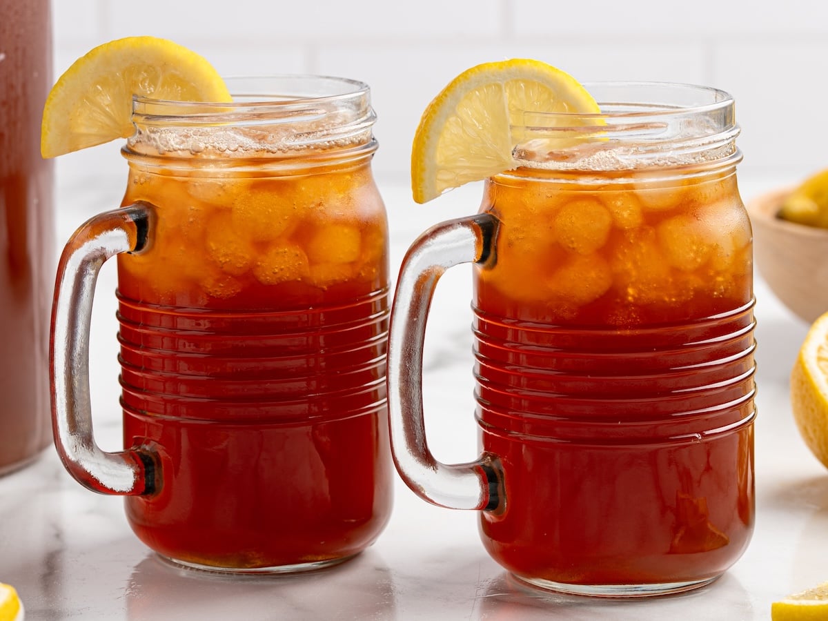 Two mason jar glasses filled with a refreshing Iced Tea recipe, packed with ice cubes and garnished with lemon wedges on the rim, are placed on a white surface.