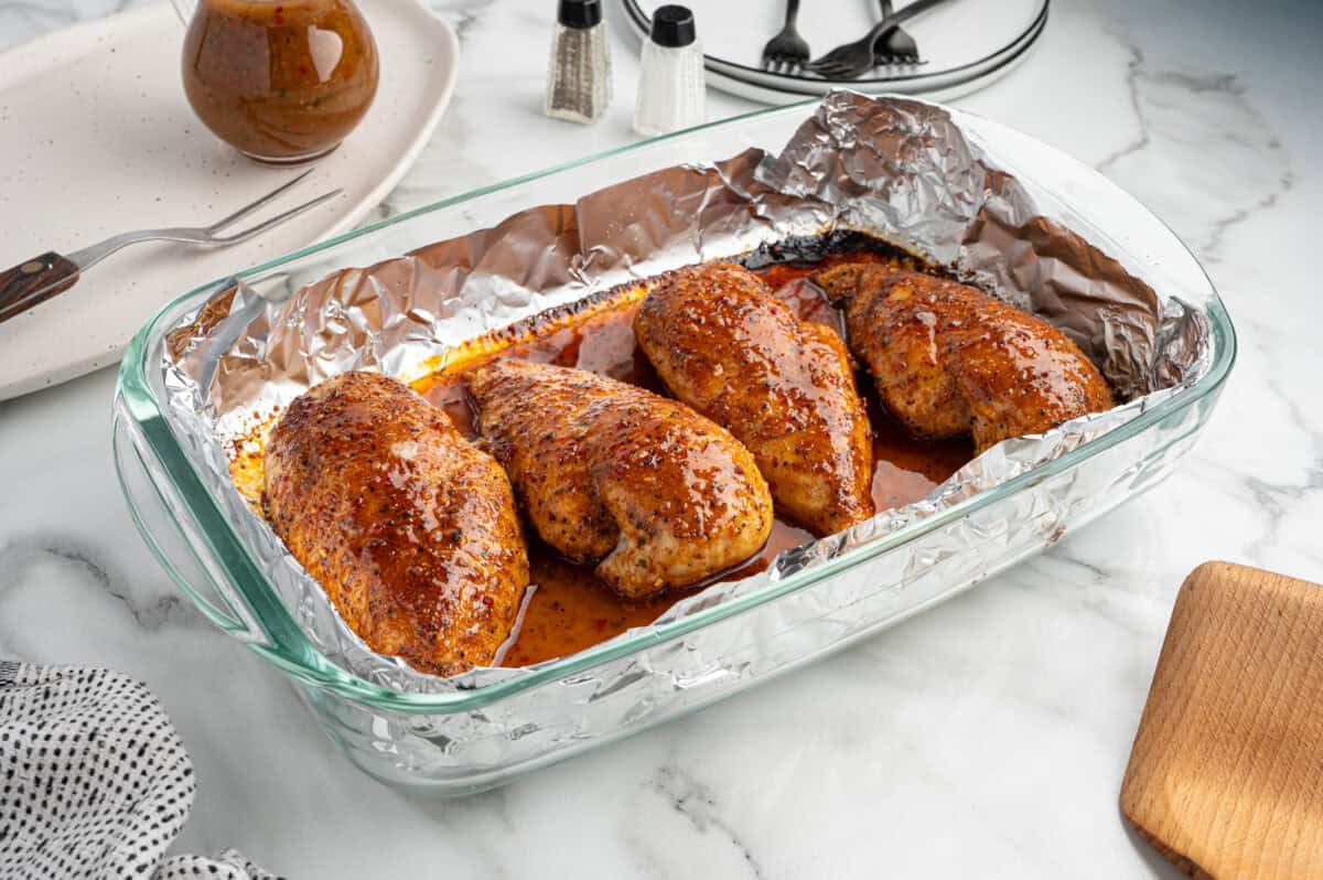 Four seasoned, baked chicken breasts in a glass dish lined with aluminum foil, sitting on a marble countertop with utensils, a napkin, and condiments nearby—an inviting presentation for this classic Italian Chicken Recipe.