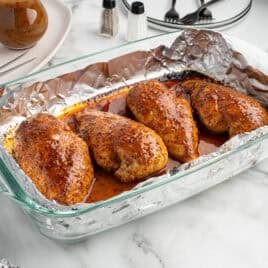 Four seasoned, baked chicken breasts in a glass dish lined with aluminum foil, sitting on a marble countertop with utensils, a napkin, and condiments nearby—an inviting presentation for this classic Italian Chicken Recipe.
