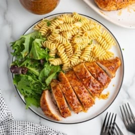 A plate with sliced grilled chicken breast, rotini pasta, and mixed greens on a marble surface; a fork and a napkin are next to the plate—an inviting presentation ideal for showcasing an Italian Chicken Recipe.