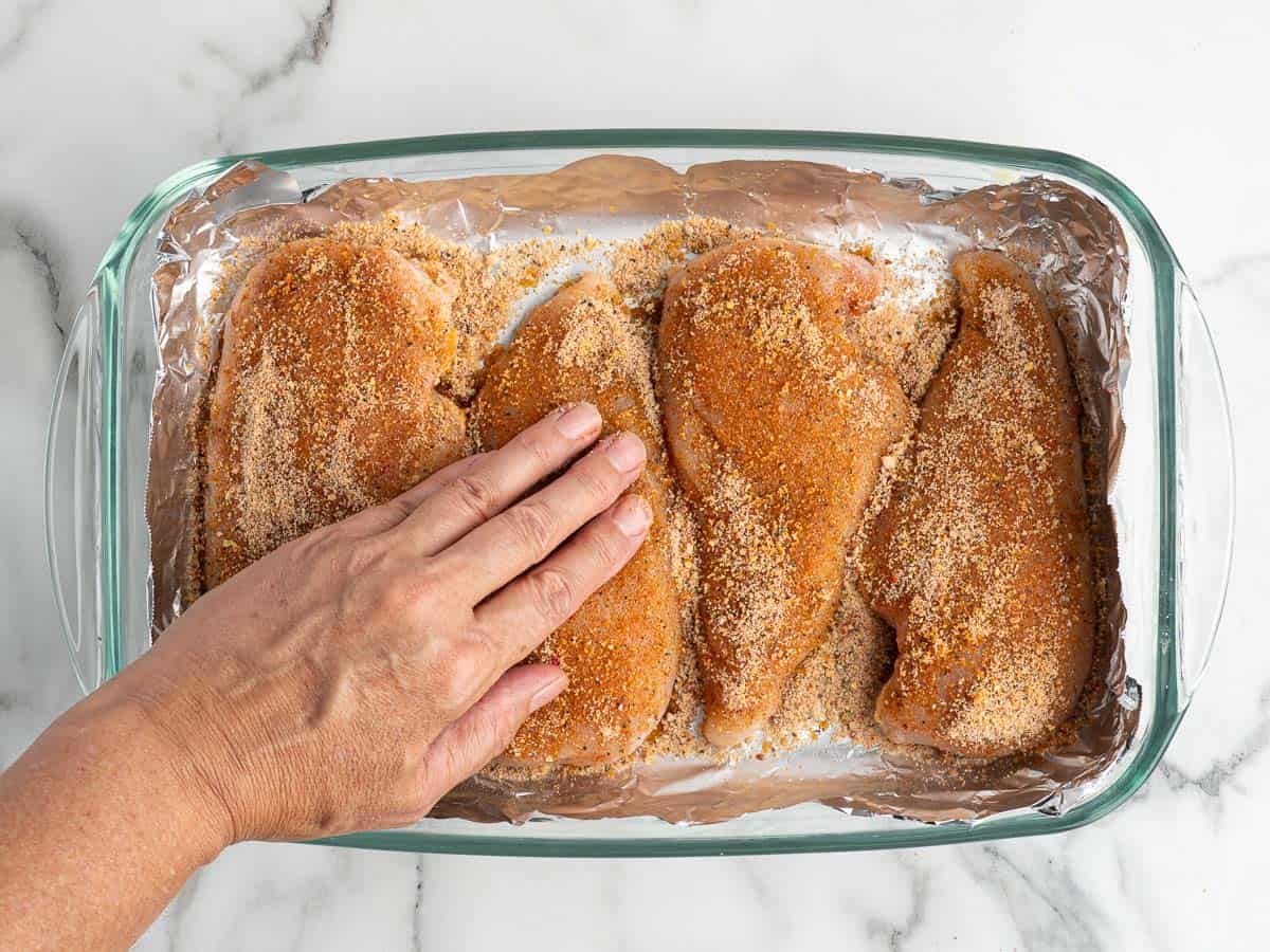 A hand presses the brown sugar seasoning onto chicken breasts in a foil-lined glass baking dish on a white marble surface.