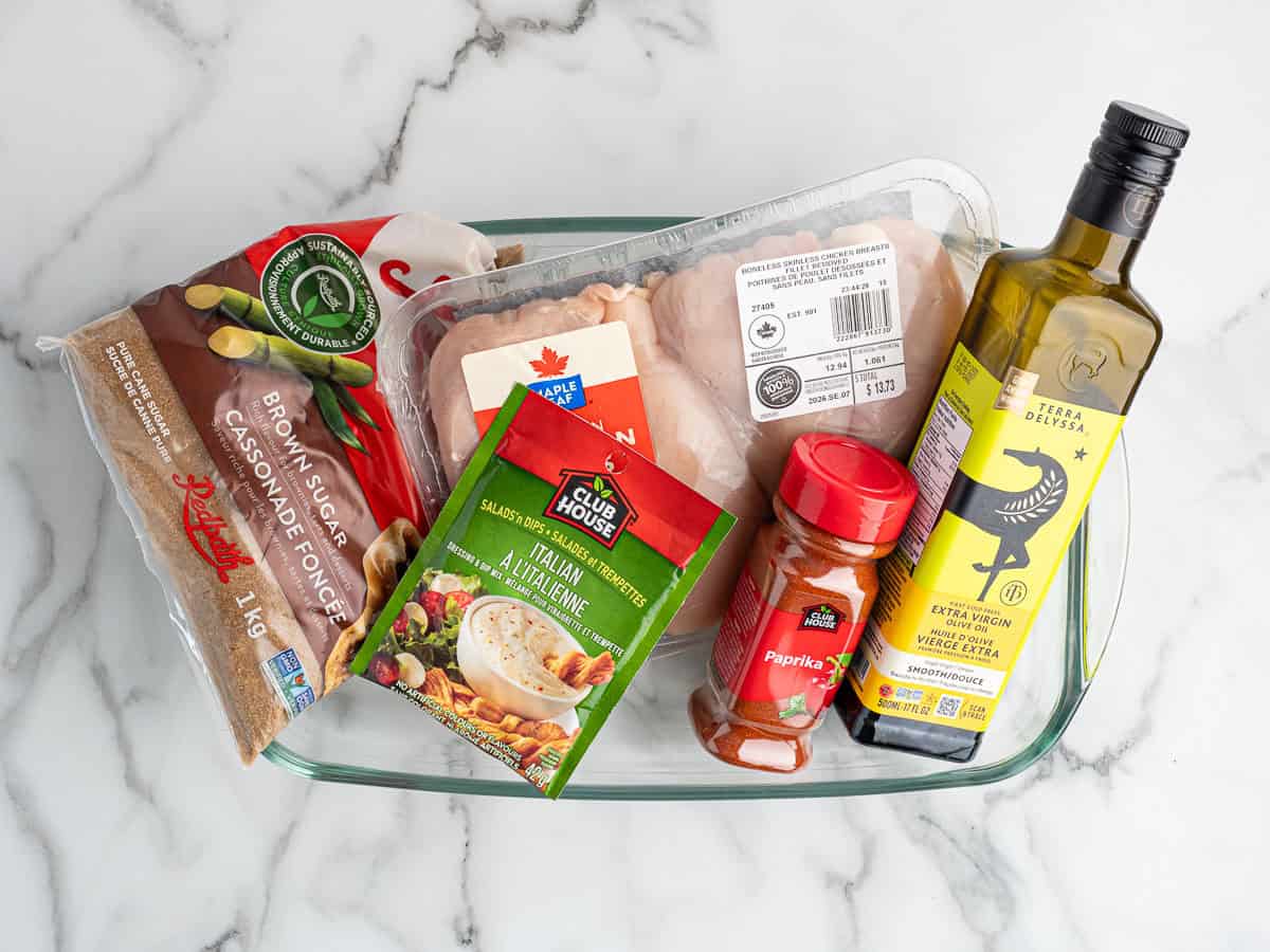 A glass dish of boneless skinless chicken breasts on a marble surface with the other ingredients for brown sugar Italian chicken: light brown sugar, a packet of dry Italian dressing mix, sweet paprika and olive oil.