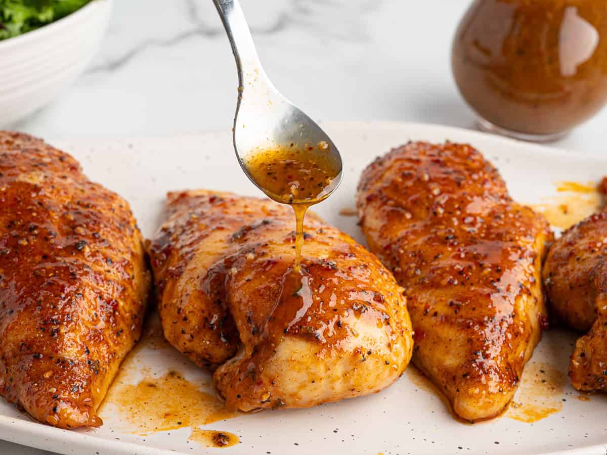 A spoon drizzles sauce over seasoned, baked chicken breasts on a white plate in this Italian Chicken Recipe, with a bowl of greens and a jar of extra sauce in the background.