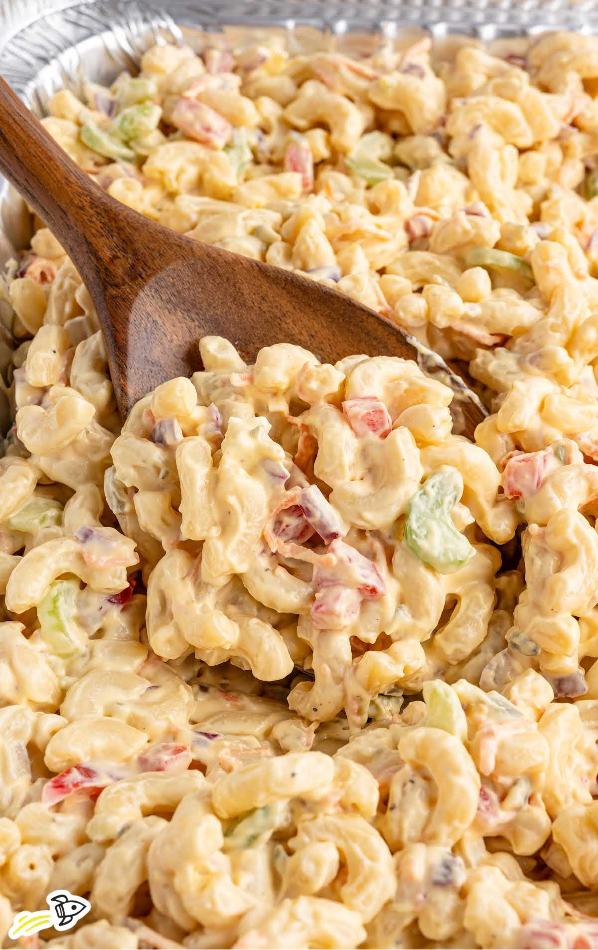 Macaroni salad in a foil tray being scooped with a wooden spoon, showcasing the creamy texture and colorful mix-ins.
