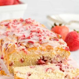 a loaf of strawberry bread with strawberries and glaze on top with a slice cut off on a wooden board