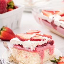close up shot of a slice of strawberry poke cake topped with whipped cream and strawberry slices on a plate