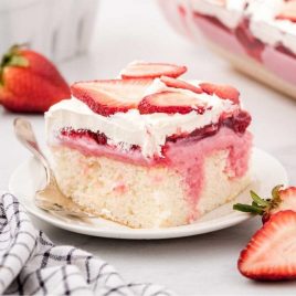 close up shot of a slice of strawberry poke cake topped with whipped cream and strawberry slices on a plate