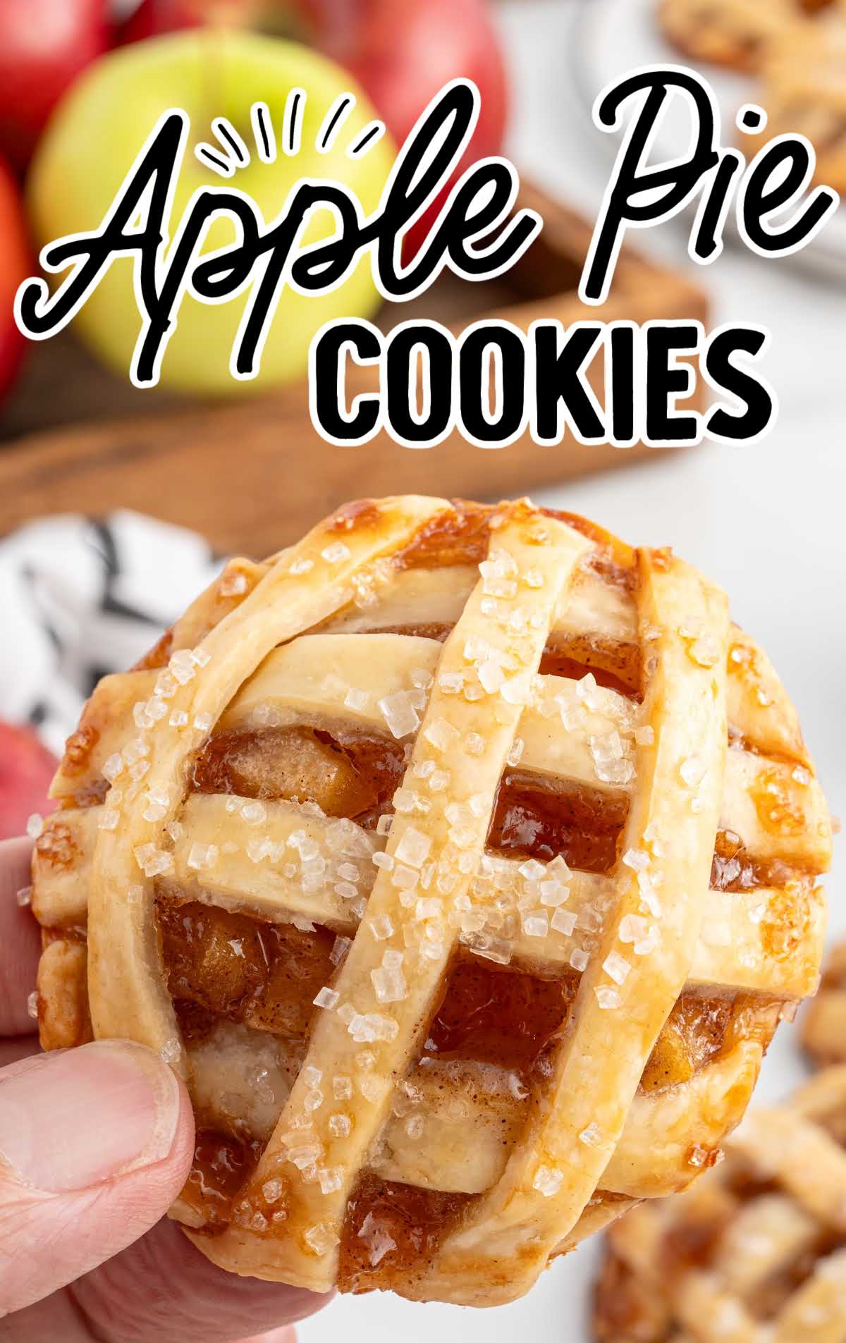 Hand holding a single apple pie cookie in front of a background of apples and more cookies.