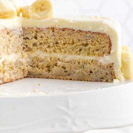 close up shot of a Banana Cake topped topped with slices of banana with a slice missing in a cake stand