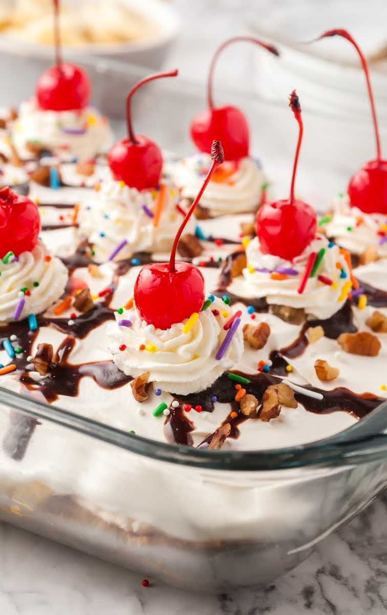 close up shot of a slice of banana split cake topped with chocolate syrup, sprinkles, whipped cream, and a cherry in a baking dish