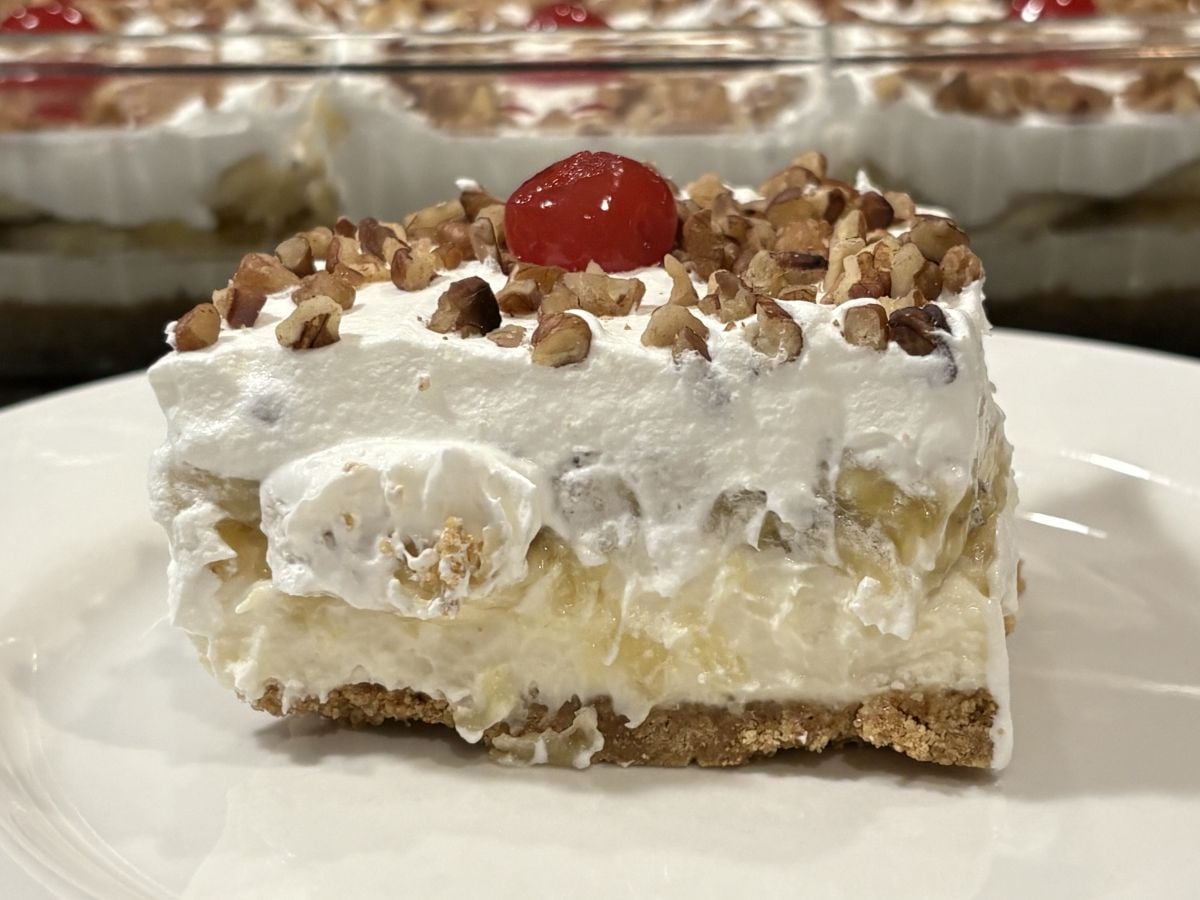 A slice of Banana Split Cake with a graham cracker crust, creamy filling, pineapple chunks, whipped topping, chopped pecans, and a cherry on top is served on a white plate. The rest of the Banana Split Cake is visible in the background.