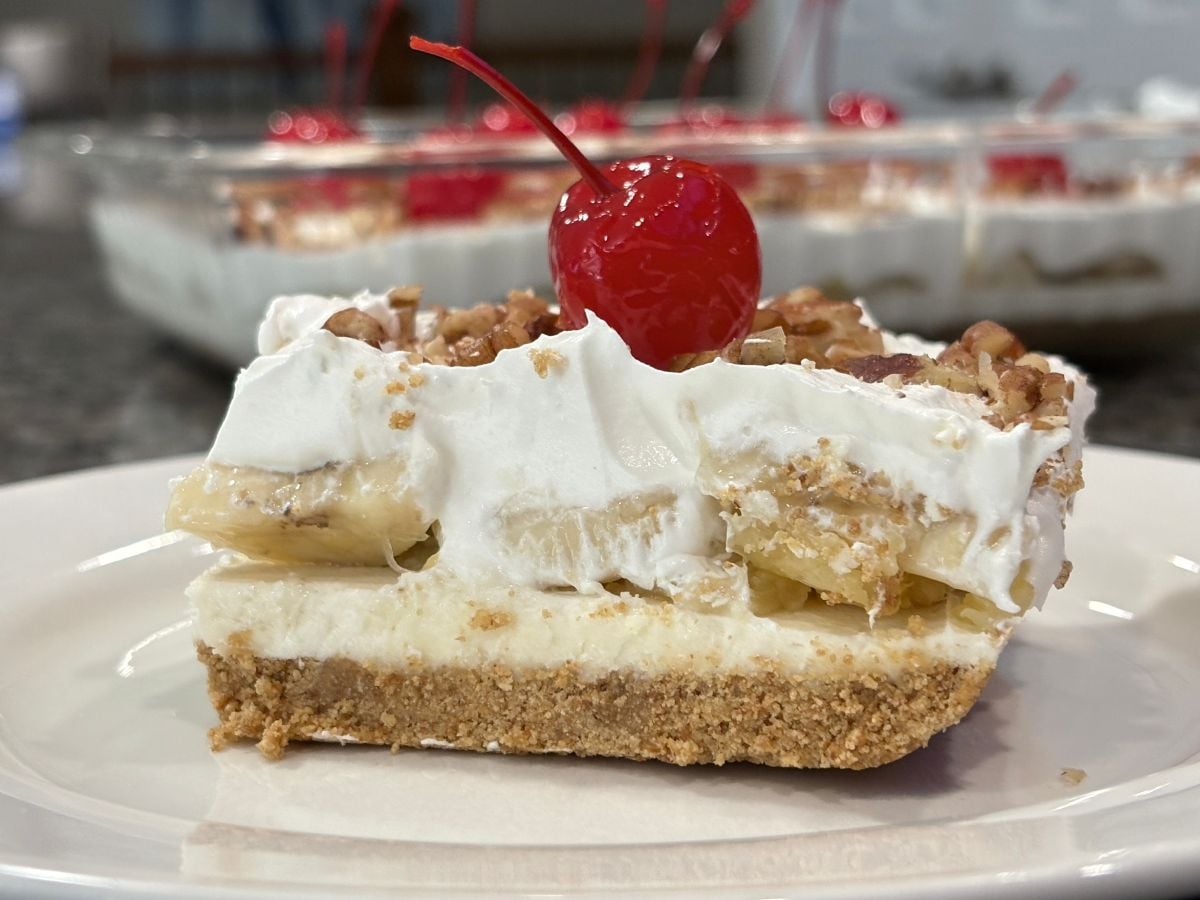 A slice of Banana Split Cake with a graham cracker crust, creamy filling, sliced bananas, whipped topping, chopped pecans, and a maraschino cherry on top, served on a white plate.