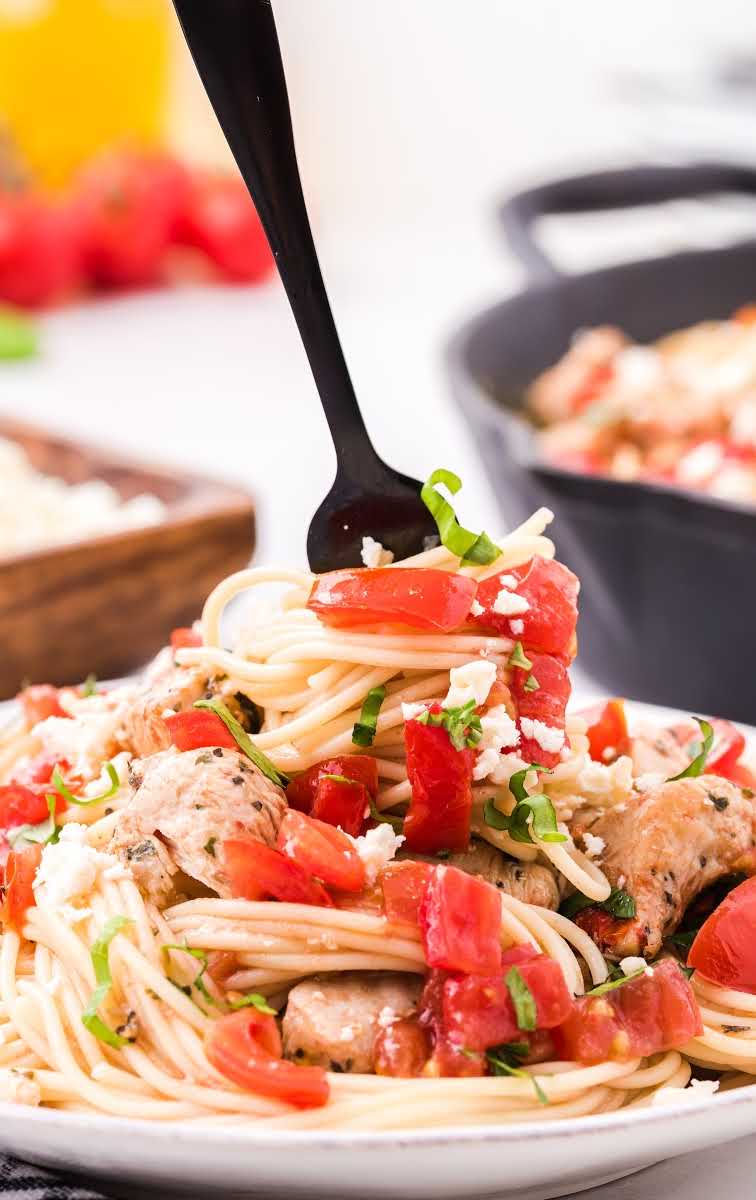 close up shot of bruschetta chicken pasta topped with parsley and feta cheese on a plate with pasta wrapped around a fork