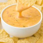 close up shot of a bowl of Cheese Dip with a tortilla chip being dipped into it