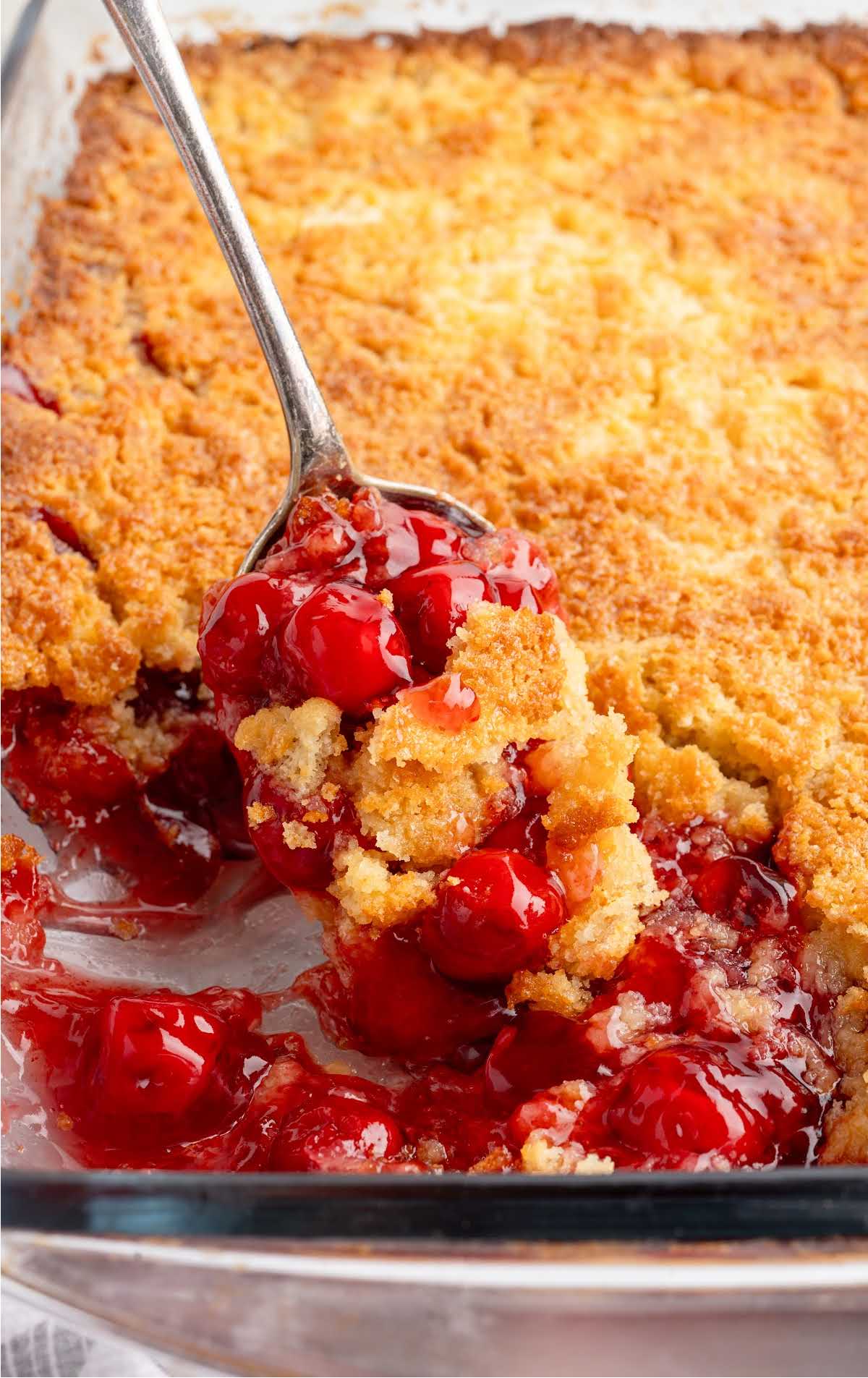 Spoonful of cherry dump cake being served from a glass baking dish with a crisp golden topping.