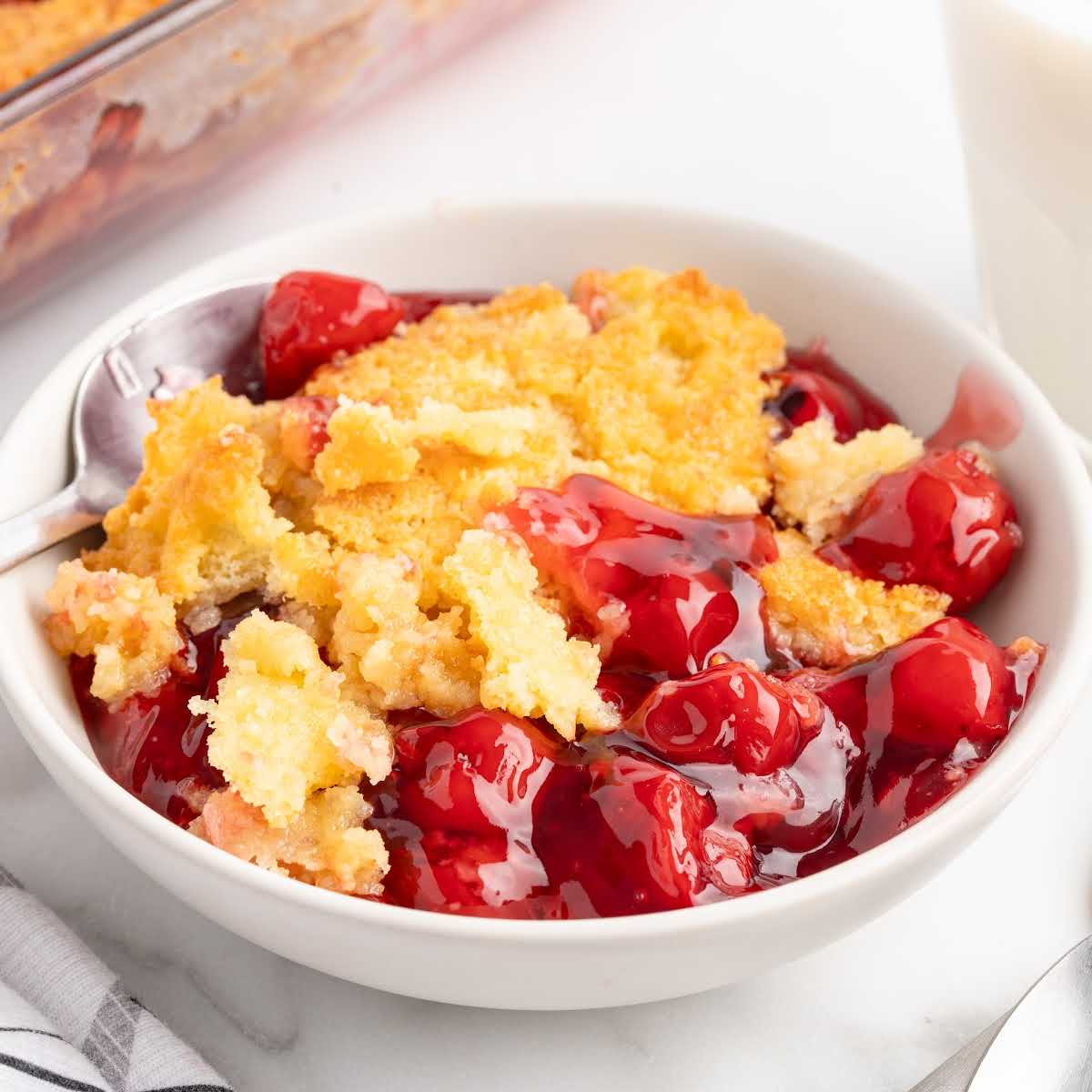 Bowl of cherry dump cake with a spoonful served beside the baking dish, showing golden crust and cherry filling.