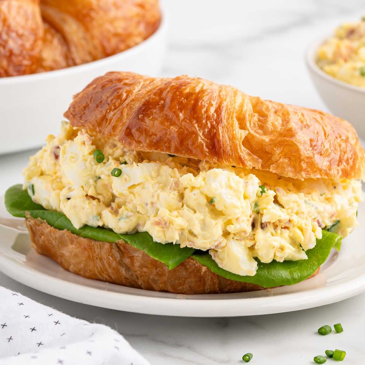 Classic egg salad sandwich on a croissant with fresh lettuce on a white plate, bowl of filling in the background.