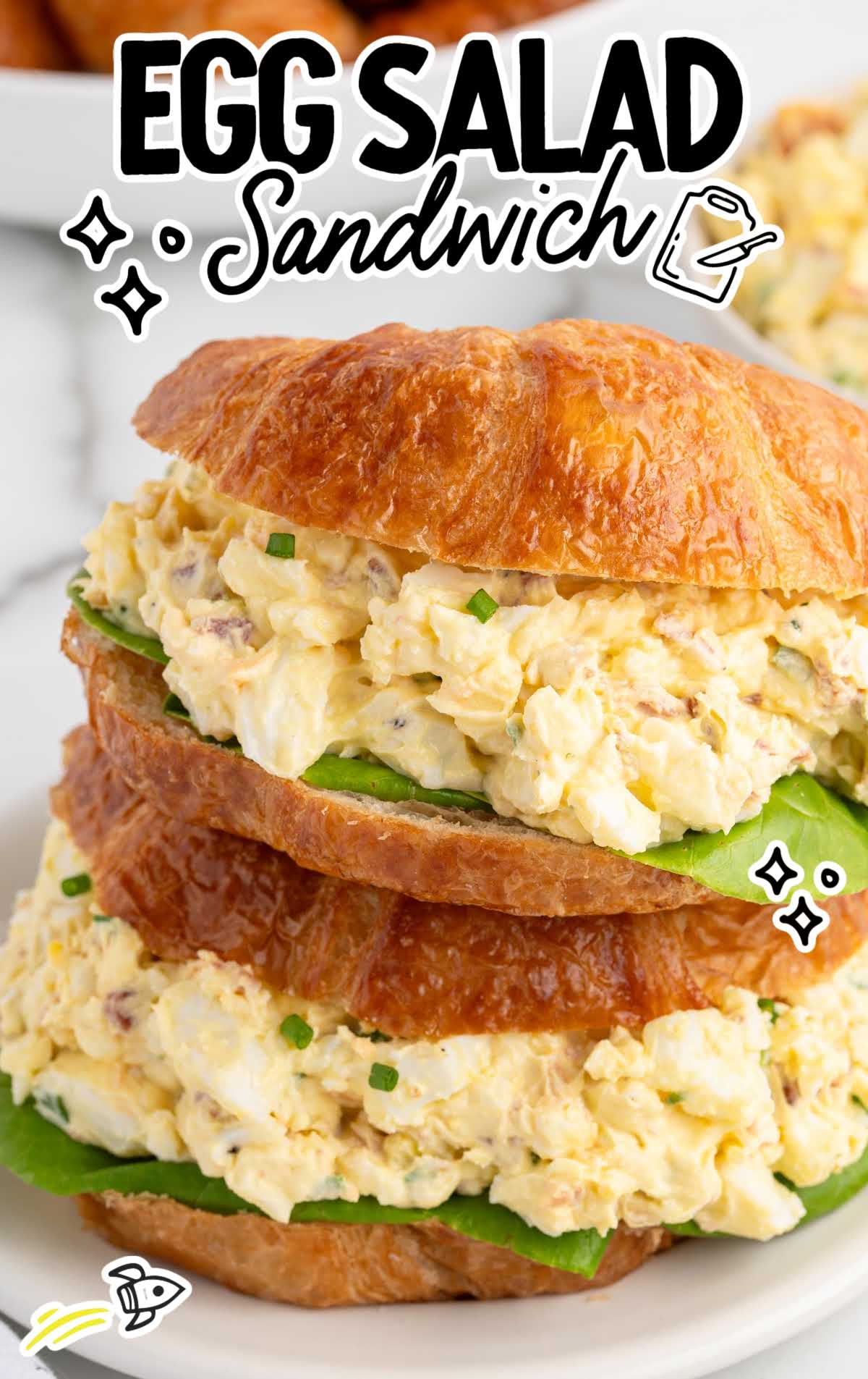 Croissant sandwich with creamy egg salad and green lettuce on a plate, surrounded by bowls of ingredients.