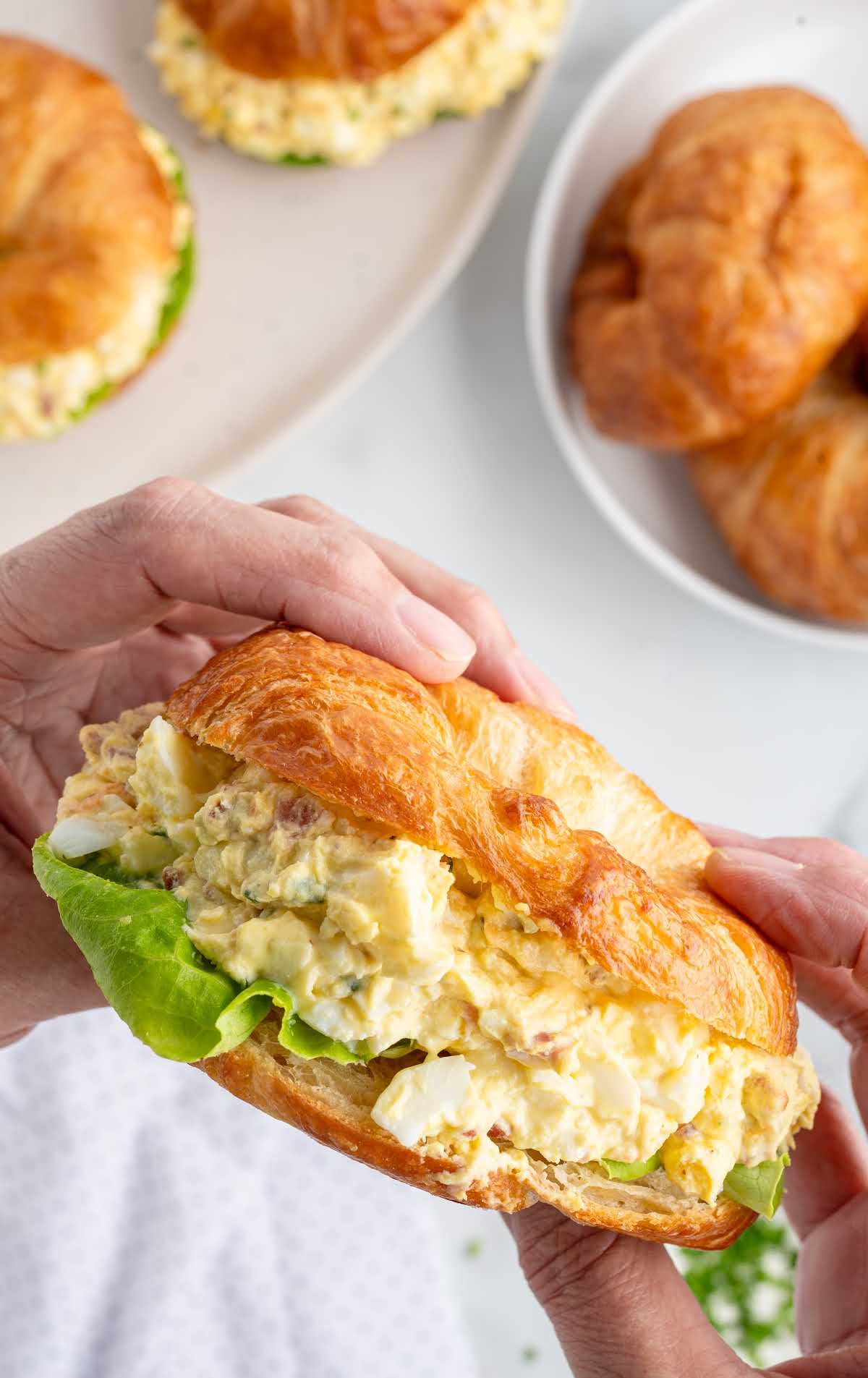 Hands holding a croissant sandwich filled with egg salad, with plates of sandwiches in the background.