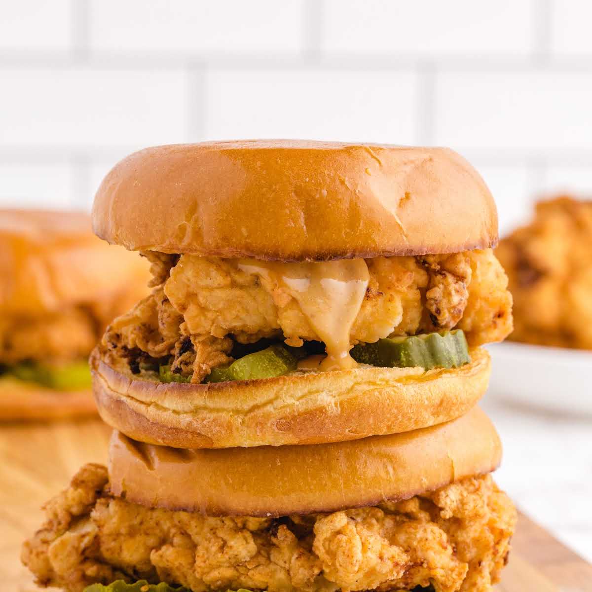 Stacked fried chicken sandwich with pickles and sauce on a wooden surface, surrounded by more sandwiches in the background