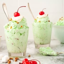Two glasses filled with light green pistachio salad, topped with whipped cream, chopped nuts, and a cherry, sit on a marble surface with extra cherries, marshmallows, and a bowl of Watergate Salad in the background.