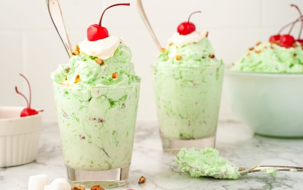 Two glasses filled with light green pistachio salad, topped with whipped cream, chopped nuts, and a cherry, sit on a marble surface with extra cherries, marshmallows, and a bowl of Watergate Salad in the background.