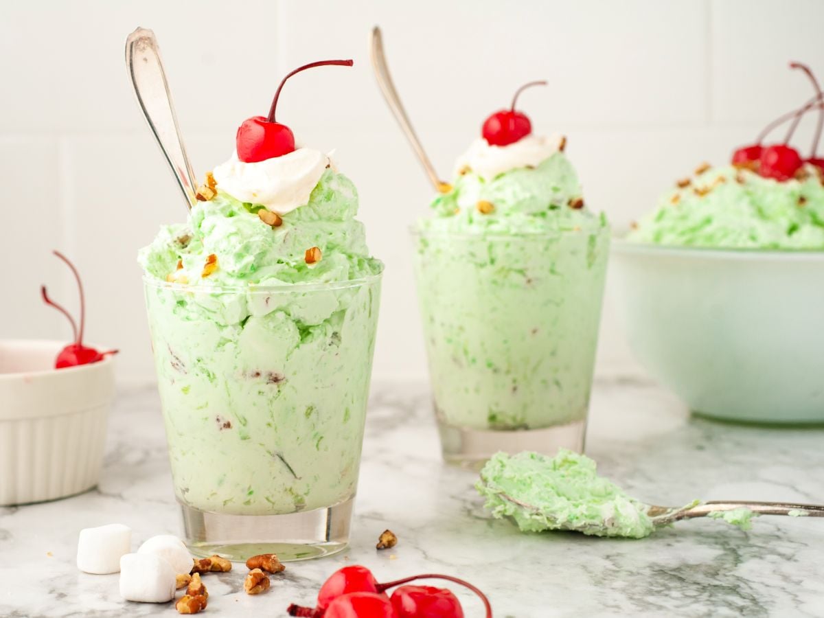 Two glasses filled with light green pistachio salad, topped with whipped cream, chopped nuts, and a cherry, sit on a marble surface with extra cherries, marshmallows, and a bowl of Watergate Salad in the background.