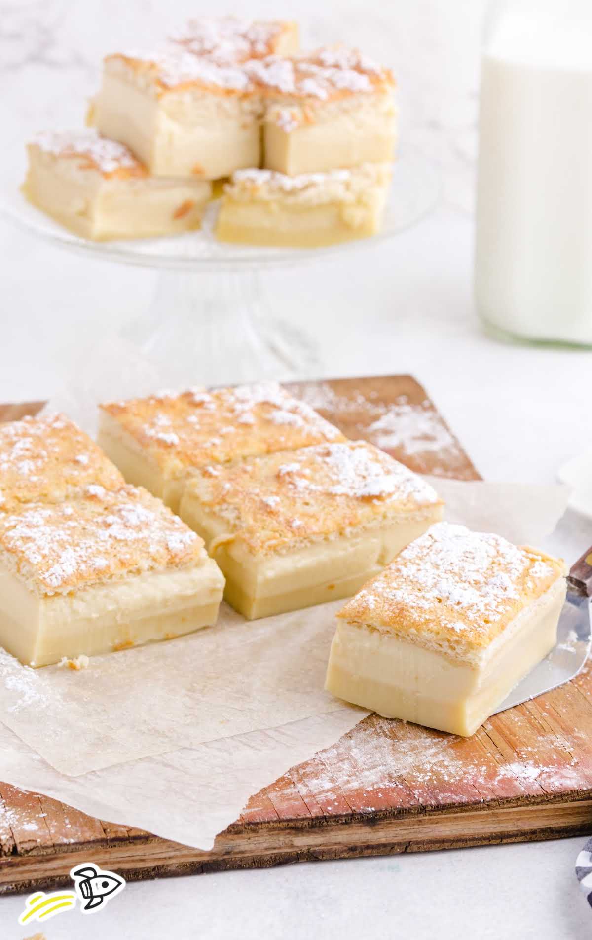 Serving board with multiple squares of magic custard cake and a glass cake stand of extras in the background.