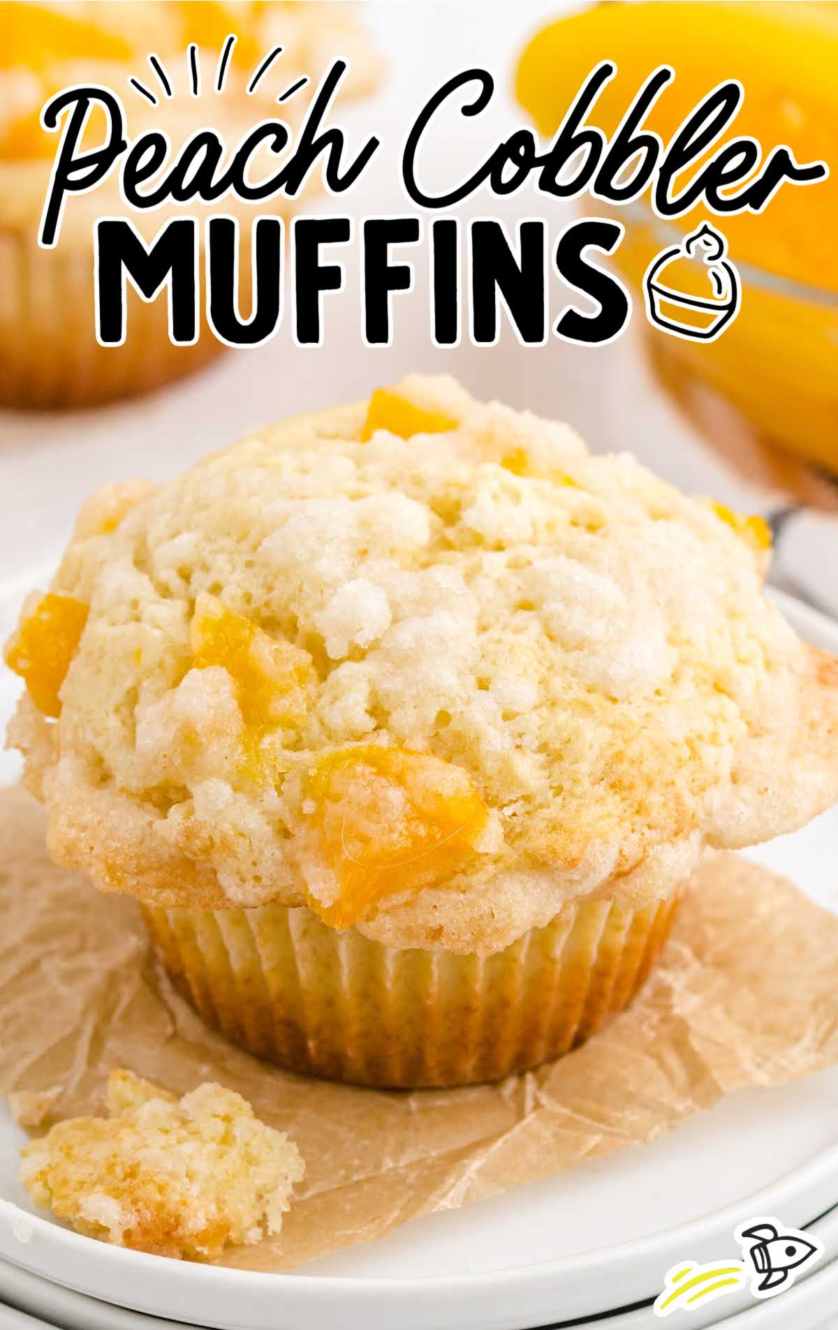 Close-up of a peach cobbler muffin in a wrapper on parchment paper, showing golden streusel topping and peach bits.