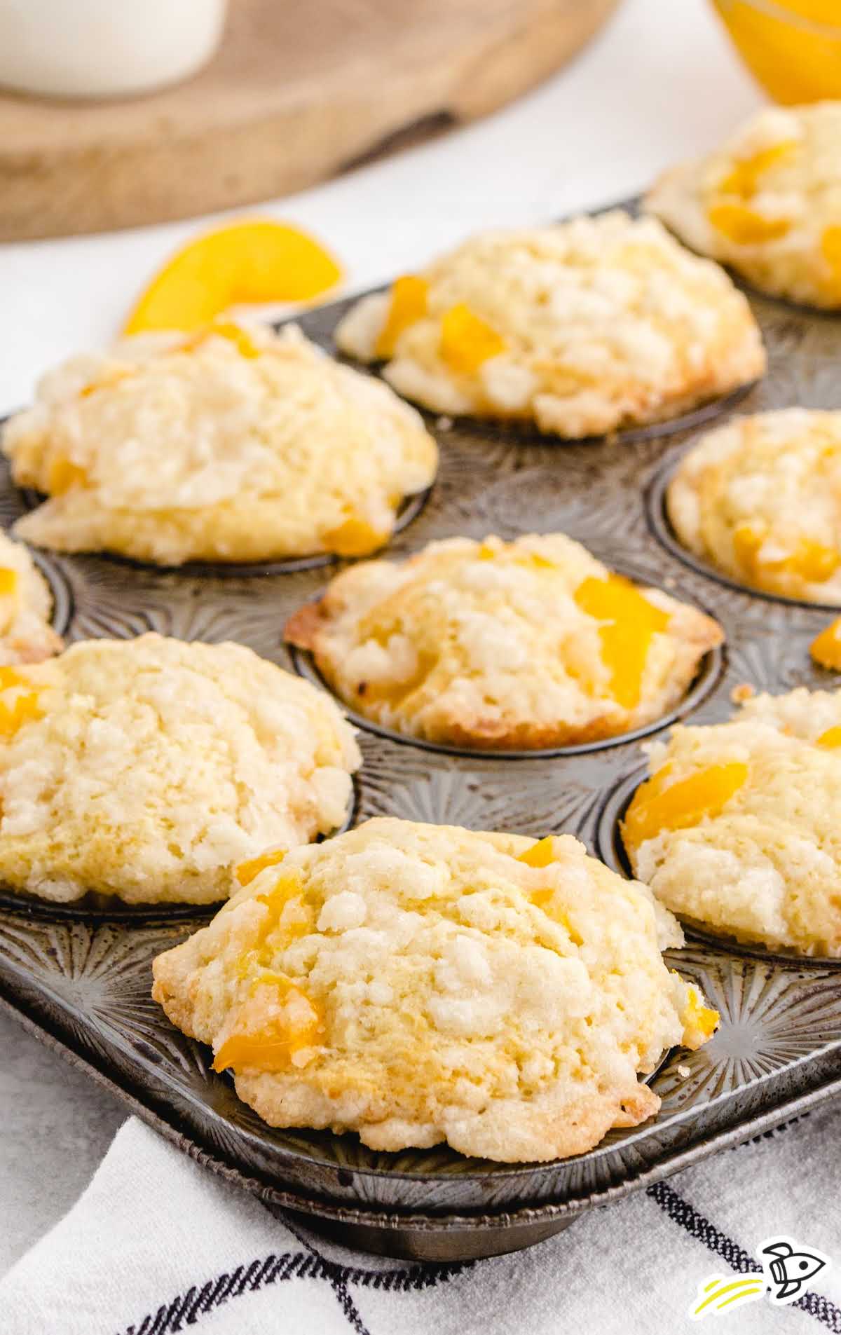 Baked peach cobbler muffins in a dark muffin tin, topped with coarse sugar and juicy peach chunks.