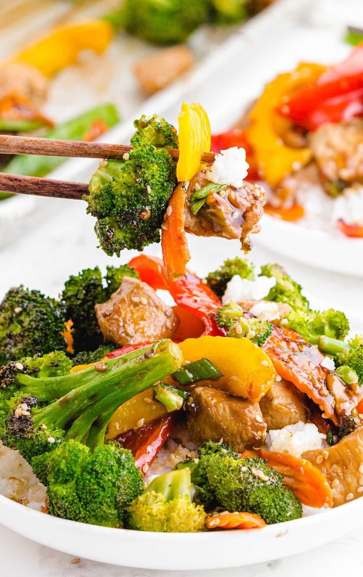 Close-up of a bowl of stir fry with broccoli, bell peppers, chicken, and rice, with chopsticks lifting a bite.