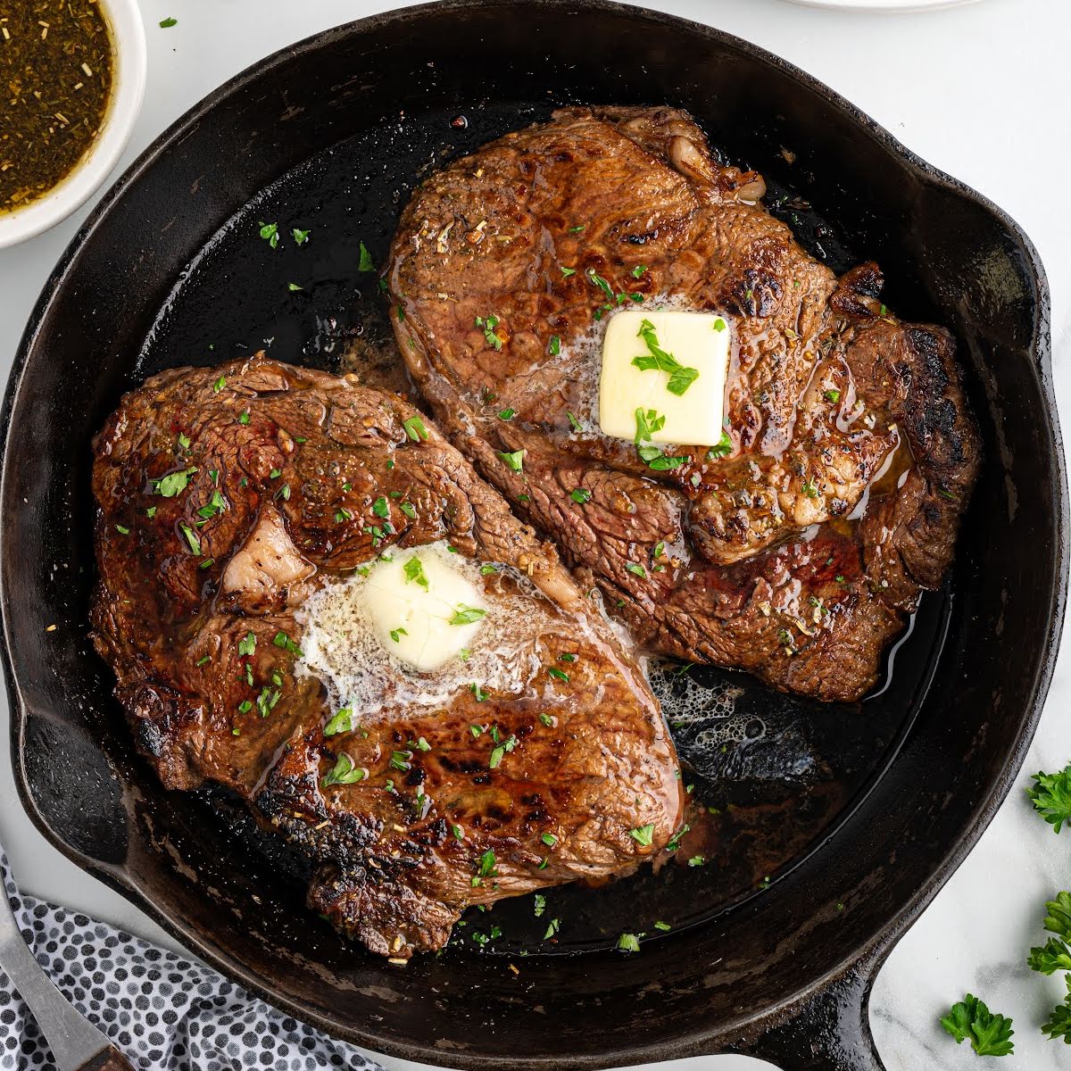 close up shot of pieces of steak topped with slices of butter and garnished with parsley in a skillet