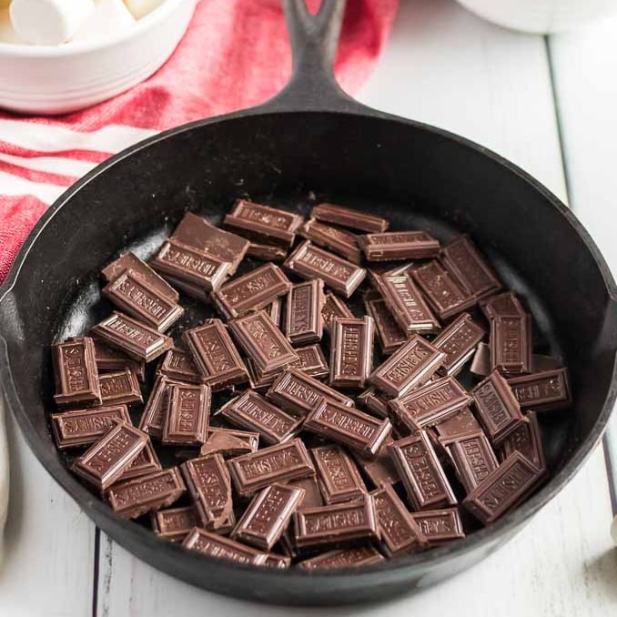 A black cast iron skillet filled with small, unwrapped Hershey&rsquo;s chocolate bar pieces sits on a white wooden surface near a red cloth and a bowl of marshmallows, ready for the perfect S'mores Dip Recipe.
