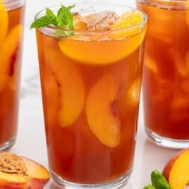 Close-up of three glasses of peach iced tea with fresh peach slices and mint leaves, surrounded by whole and cut peaches.