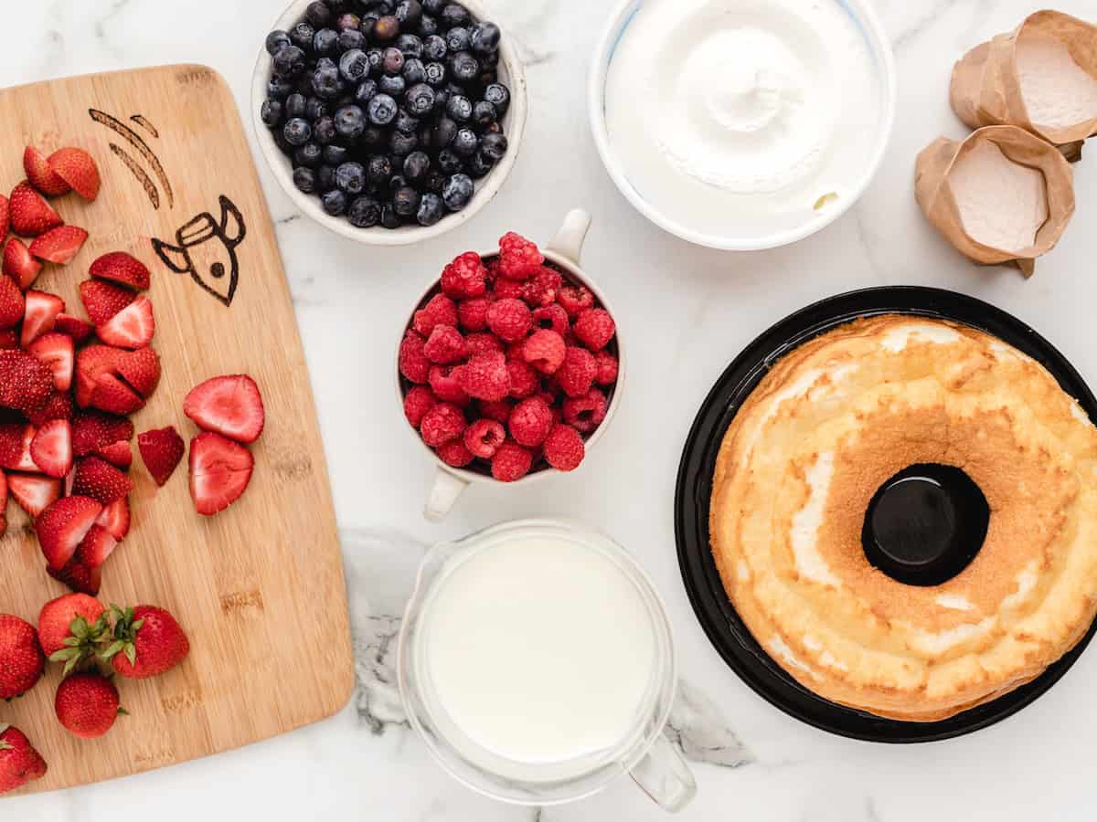 A round angel food cake, sliced strawberries on a cutting board, bowls of blueberries, raspberries, and whipped cream, plus a jug of milk and two cracked eggshells on a marble surface—perfect ingredients for creating a classic Vanilla Trifle recipe.