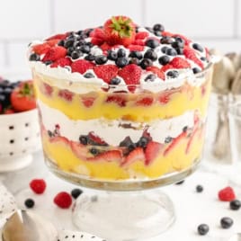 A classic Vanilla Trifle recipe, this layered dessert features whipped cream, custard, and mixed berries beautifully arranged in a glass bowl. Strawberries, blueberries, and raspberries decorate the top of this irresistible vanilla trifle. In the background, a bowl of berries and spoons await for serving.