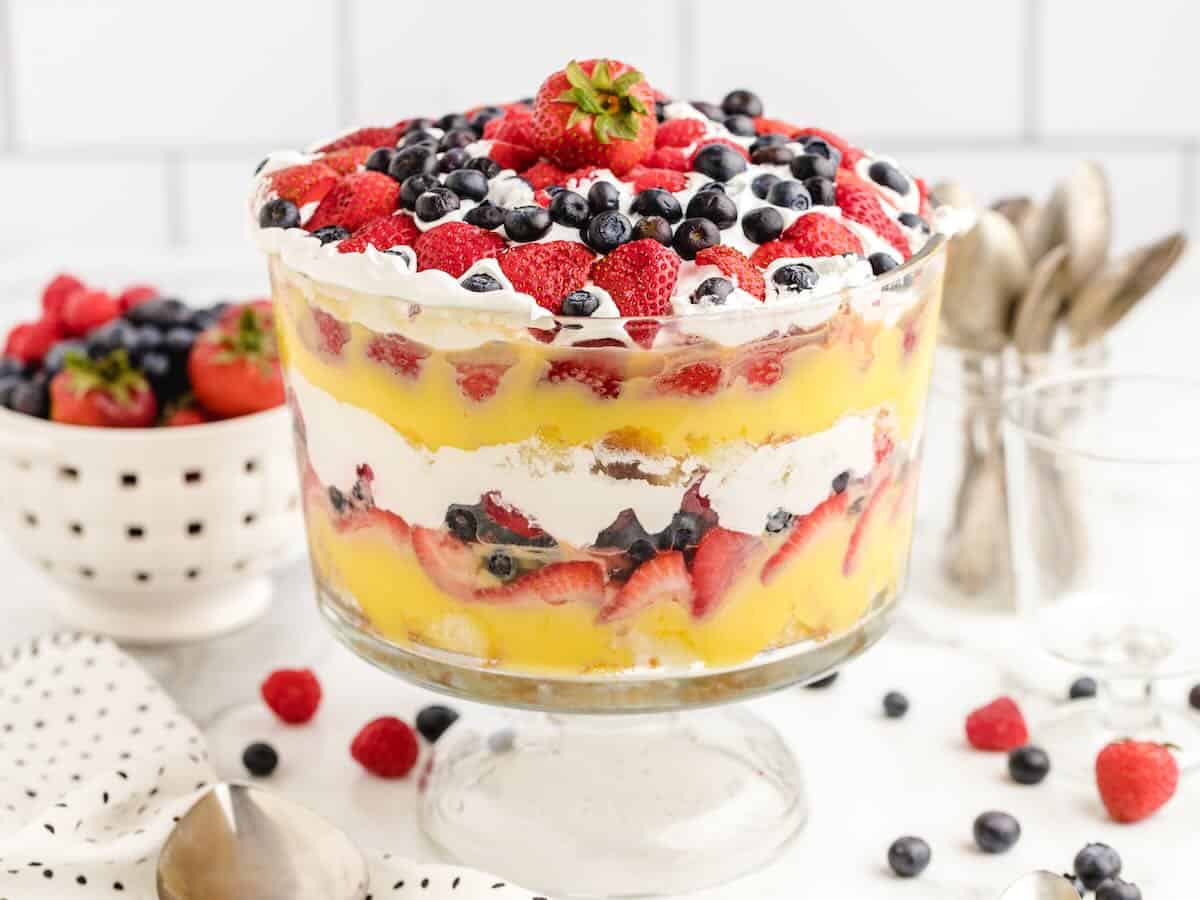 A classic Vanilla Trifle recipe, this layered dessert features whipped cream, custard, and mixed berries beautifully arranged in a glass bowl. Strawberries, blueberries, and raspberries decorate the top of this irresistible vanilla trifle. In the background, a bowl of berries and spoons await for serving.